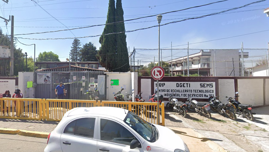 Fotografía en la que se muestra la entrada del plantel, conformada por una reja de color gris que da acceso a las instalaciones. Del lado derecho de la reja hay un muro de color blanco que tiene un borde rojo. Sobre el muro está escrito el nombre del plantel.